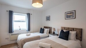 Habitación de hotel con dos camas, una ventana con cortinas, un radiador, un cuadro en la pared y una lámpara en el techo.