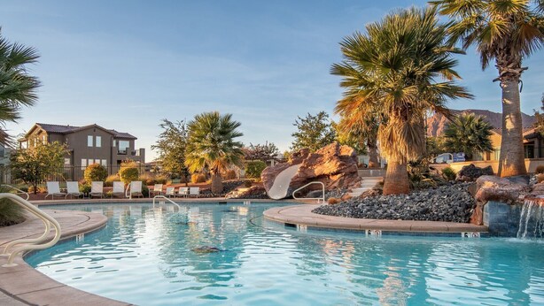 A swimming pool with a waterfall feature, surrounded by palm trees and a building.