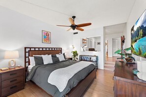 A bedroom with a bed, nightstands, a ceiling fan, a bedside table with a lamp, a bookshelf, and a large painting on the wall. - New Family Retreat Near Beaches & Whalers Village (Lahaina)