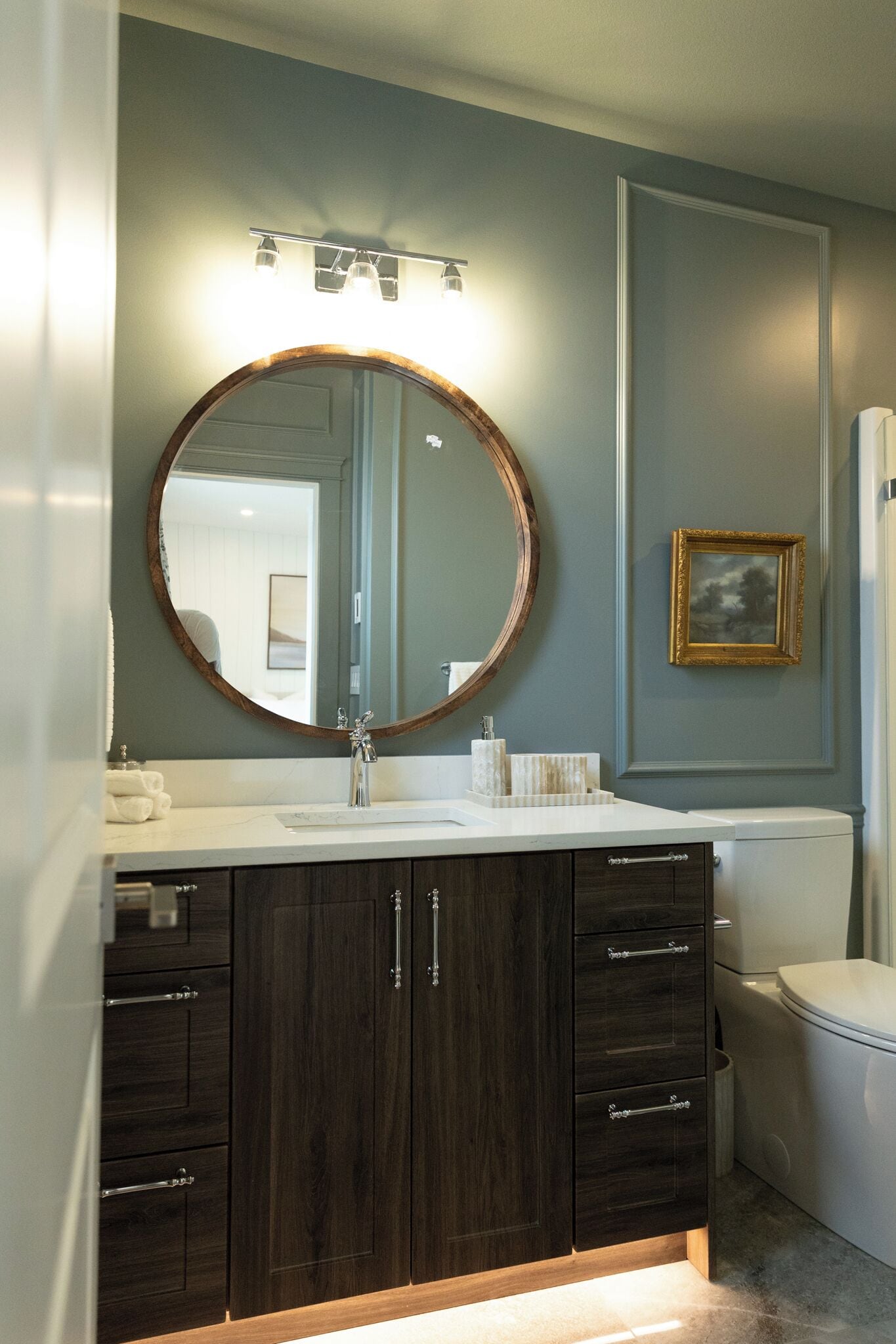 A bathroom with a dark wood vanity, a round mirror, a white sink, and a toilet.