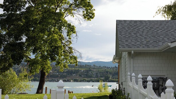 Una casa con cerca blanca, un árbol grande y vista a un lago con montañas al fondo.