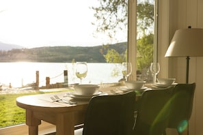 A dining table with white plates, wine glasses, and cutlery set for two, overlooking a lake with a bridge in the background.