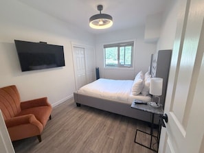 Ein Schlafzimmer mit einem Bett, einem Sessel, einem Schreibtisch mit Lampe, einem Fernseher und einem Fenster.