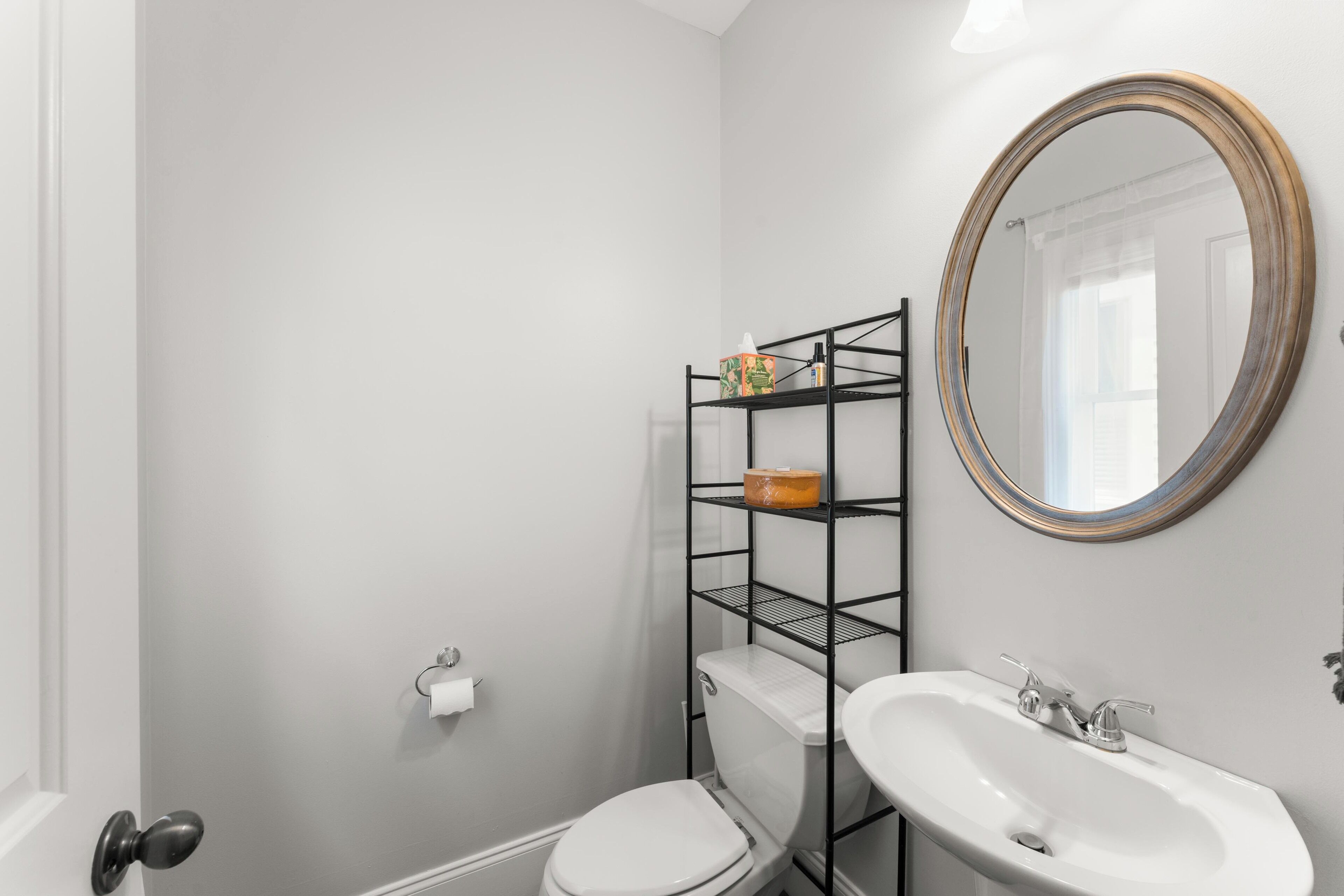 Un baño con inodoro y lavamanos blancos, estante metálico negro con artículos de aseo, y un espejo redondo en la pared.