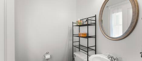 Un baño con inodoro y lavamanos blancos, estante metálico negro con artículos de aseo, y un espejo redondo en la pared.