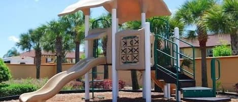 A playground with a slide and climbing structure, surrounded by palm trees and a clear sky.