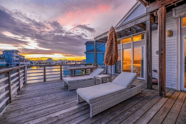 A wooden deck with lounge chairs and a thatched umbrella, overlooking a body of water and buildings during sunset.