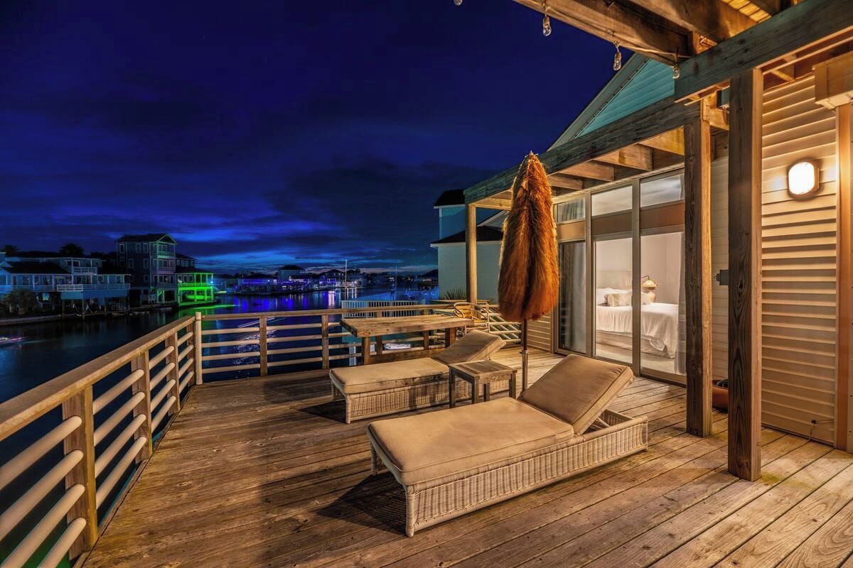 Une terrasse en bois avec des meubles en osier, une porte vitrée donnant sur une pièce et une vue sur une marina la nuit.