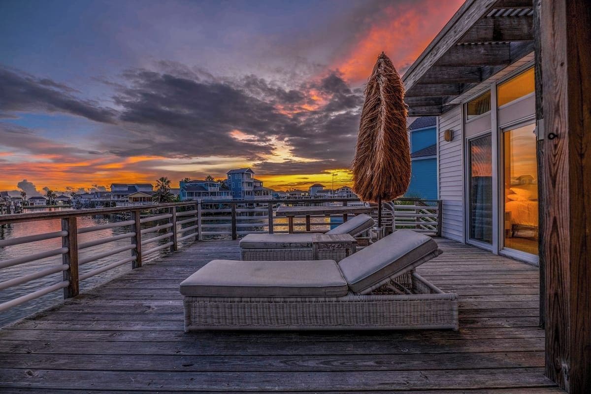 A wooden deck with lounge chairs and a large umbrella, overlooking a waterfront with buildings in the background.