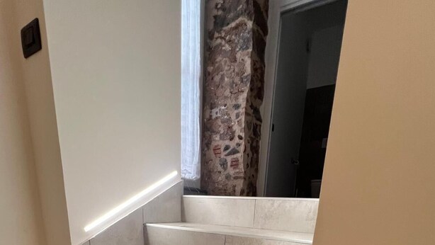 A set of beige tiled stairs leading to a room with a stone wall and a window with white curtains. - IN THE HEART OF CATANIA — THE PETITE SUITE SAN GIULIANO (Catania)