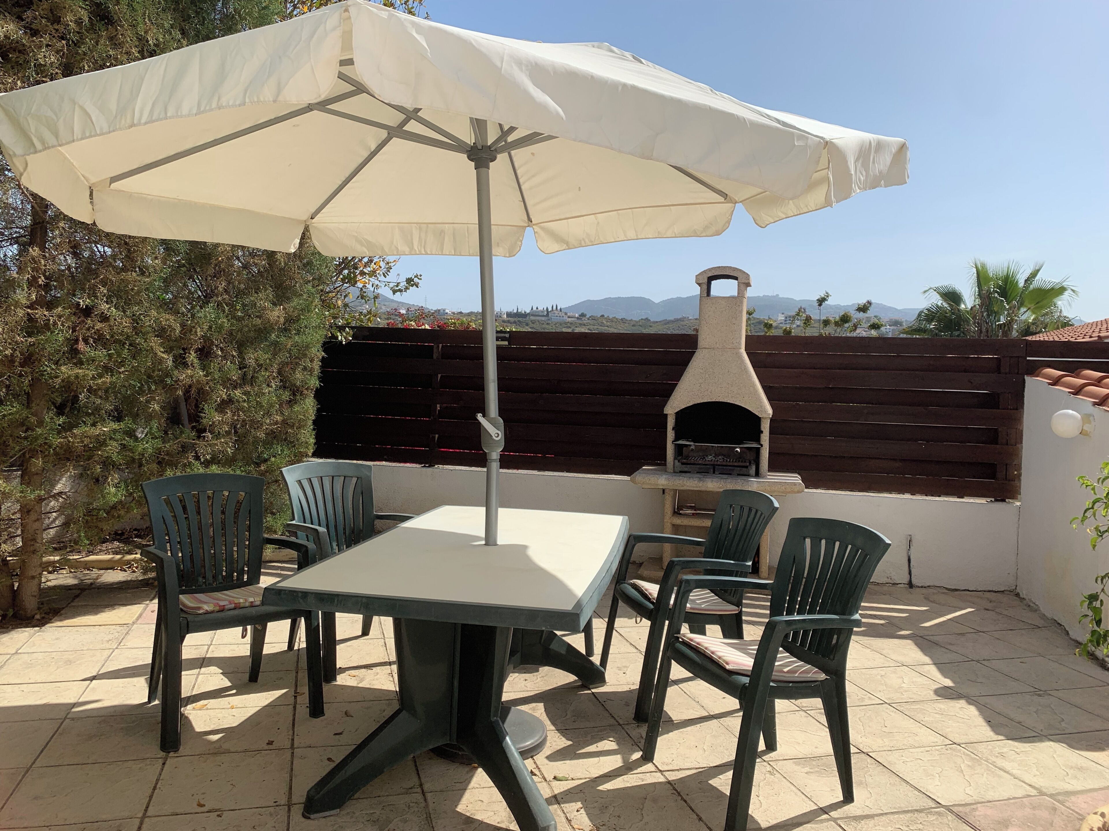 Een terras buiten met een tafel, stoelen, een witte parasol en een stenen oven.
