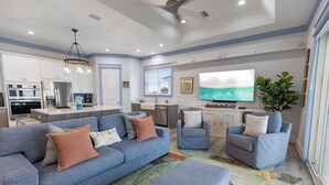 A modern kitchen with stainless steel appliances, a dining area with a blue sofa and armchairs, and a large flat-screen TV mounted on the wall. - Shore Beats Work - Gulf Views, Pool, Elevator, Beach Access (Panama City Beach)