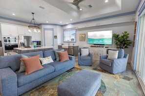 A modern kitchen with stainless steel appliances, a dining area with a blue sofa and armchairs, and a large flat-screen TV mounted on the wall. - Shore Beats Work - Gulf Views, Pool, Elevator, Beach Access (Panama City Beach)