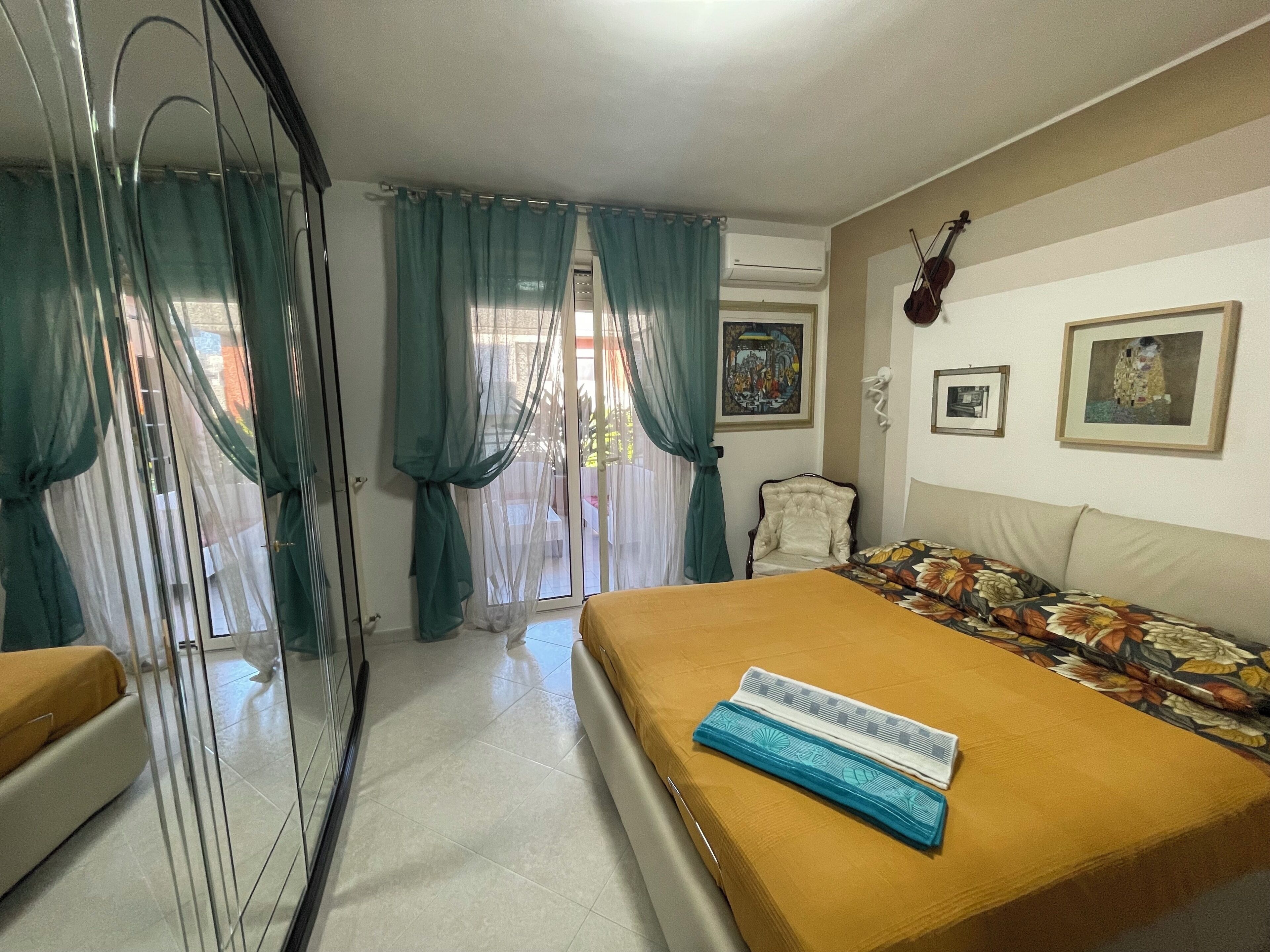 A hotel room with a bed, a chair, a violin, and a painting on the wall.