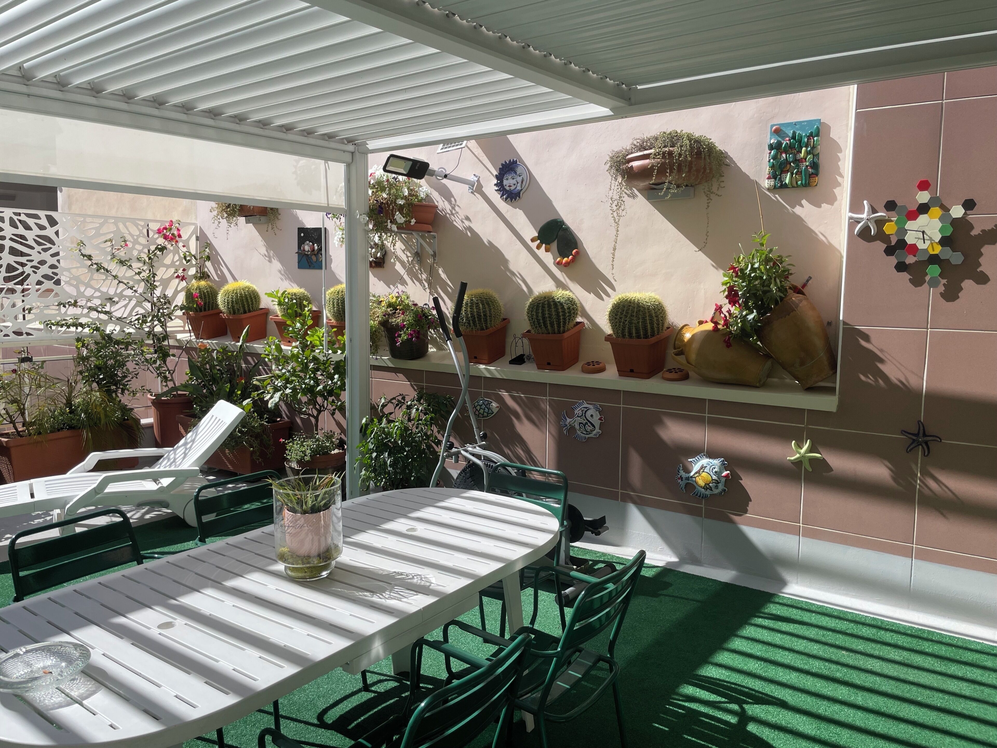 Ein überdachter Patio-Bereich mit Tisch und Stühlen, umgeben von Topfpflanzen und dekorativer Wandgestaltung.