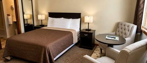 A hotel room with a large bed, two bedside tables with lamps, a round table with a chair, and a mirror on the wall.