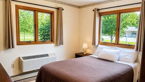 Quarto de hotel com uma cama, duas janelas com cortinas, um abajur e um aquecedor fixado na parede.
