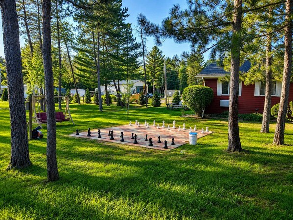 A chessboard set up on a grassy lawn surrounded by trees and a red building.
