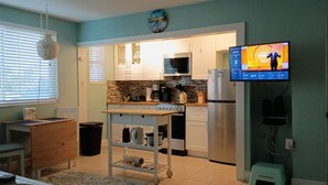 A compact kitchen with a built-in oven, microwave, and refrigerator, a small dining table, and a television displaying a news program. - The Tropical Garden Paradise🌴 on St. Pete Beach🌞⛱️🌊Friendly Native #802 (St. Pete Beach)