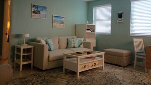 A living room with a sofa, a coffee table, a side table, a bookshelf, and a window with blinds. - The Tropical Garden Paradise🌴 on St. Pete Beach🌞⛱️🌊Friendly Native #802 (St. Pete Beach)