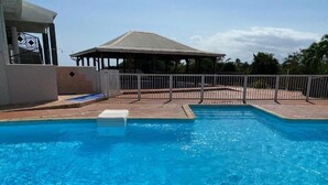 A swimming pool with a fenced deck area, a covered pavilion, and a building with a distinctive geometric design. - Brand-new studio apartment, ocean view, pool, near beaches (Le Gosier)