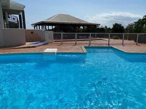 A swimming pool with a fenced deck area, a covered pavilion, and a building with a distinctive geometric design. - Brand-new studio apartment, ocean view, pool, near beaches (Le Gosier)