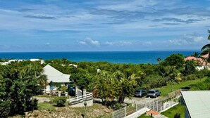 A coastal view with a white building, parked cars, and lush greenery. - Brand-new studio apartment, ocean view, pool, near beaches (Le Gosier)