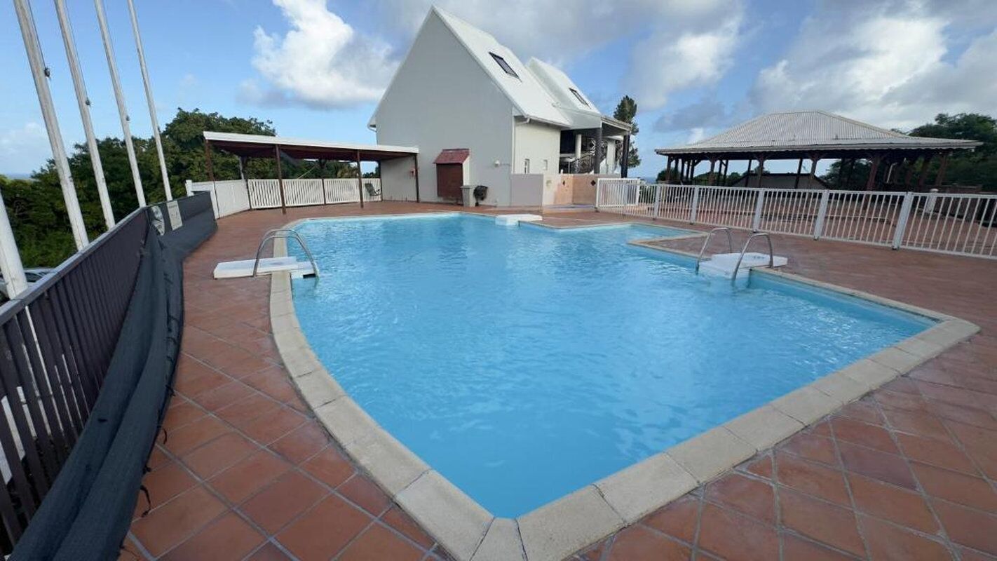 Una piscina rectangular con un borde de baldosas, un área de estar cubierta y un edificio con techo inclinado al fondo.