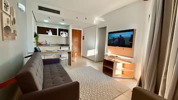 A modern living room with a sofa, a flat-screen TV mounted on a wooden stand, and a kitchen area in the background.