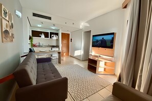 A modern living room with a sofa, a flat-screen TV mounted on a wooden stand, and a kitchen area in the background. - SHB Inigualável vista para a praia (Santos)