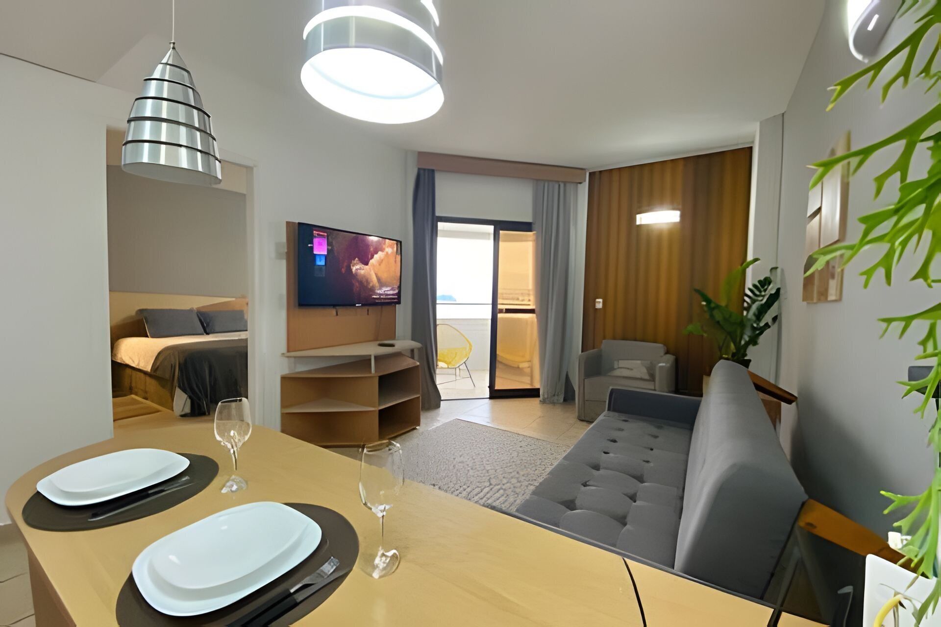 A modern living room with a dining area, a TV mounted on the wall, and a sliding door leading to a balcony.