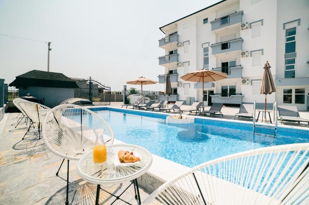 Una piscina con sillas de descanso y sombrillas, una mesa pequeña con una bebida y un edificio con balcones.