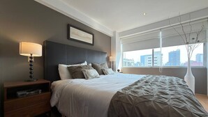 Ein modernes Schlafzimmer mit einem großen Bett, einem Nachttisch, einer Lampe und Blick auf die Stadt durch große Fenster.