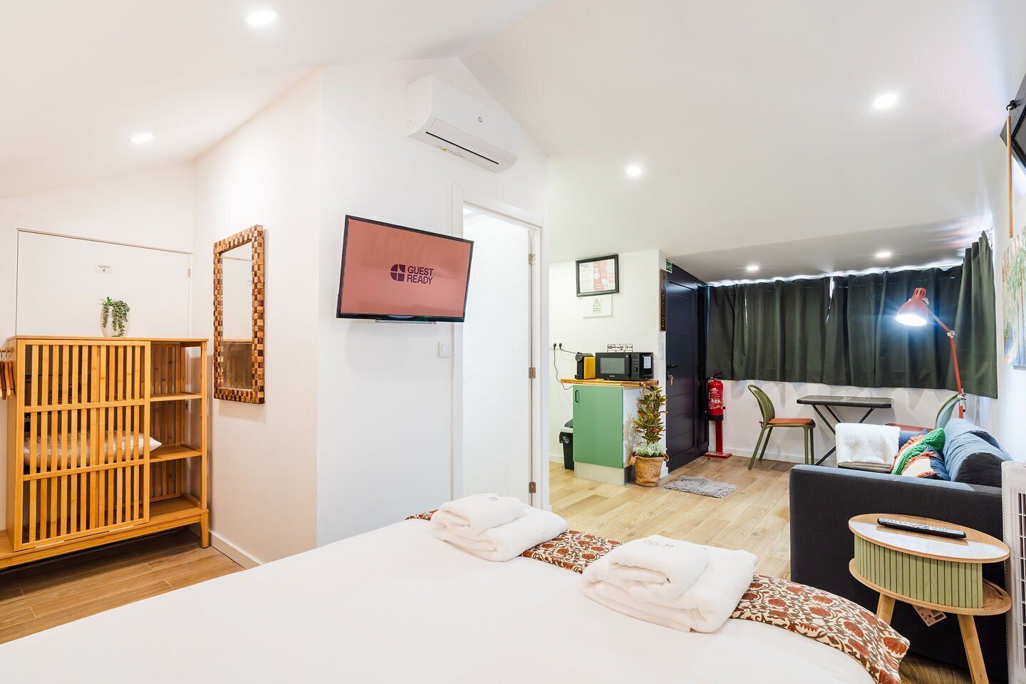 Habitación de hotel con cama, armario de madera, televisor, kitchenette, zona de comedor y ventana con cortinas.