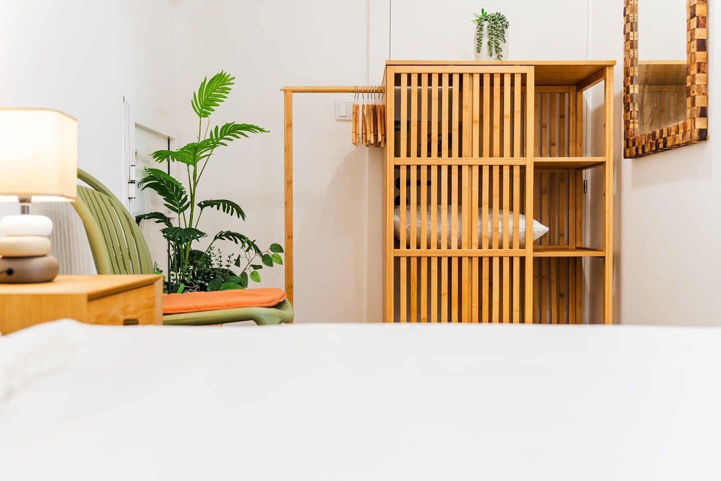 Une pièce comprenant un lit, une table de chevet avec une lampe, un fauteuil vert, une armoire en bois et une plante en pot.