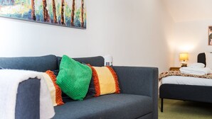 Et moderne hotelværelse med en grå sofa, farverige puder, en seng med et mønstret sengetæppe, et sengebord med en lampe og et billede på væggen.