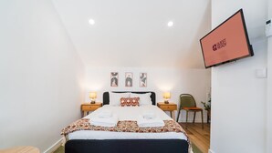 Et hotelværelse med en seng, to sengeborde med lamper, en stol, et fjernsyn og billedkunst på væggen.