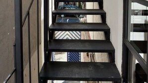 En smal, svart metalltrappa med ett glasräcke, som leder upp till en byggnad med vit fasad och utsikt över träd och parkerade bilar.