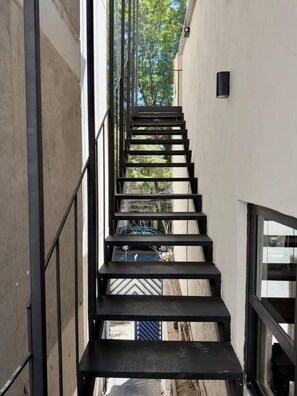 Eine schmale, schwarze Metalltreppe mit Glastrennwand, die zu einem Gebäude mit weißer Fassade und Blick auf Bäume und parkende Autos führt.