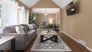 Ein Wohnzimmer mit hohen Decken, einem an der Wand befestigten Flachbildfernseher, einem grauen Sofa, einem Couchtisch und einem Teppich mit Muster.