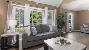 Ein Wohnzimmer mit grauem Sofa, einem Couchtisch und einem Beistelltisch mit Lampe. Große Fenster mit Vorhängen, eine Pflanze und Blick ins Grüne sind zu sehen.