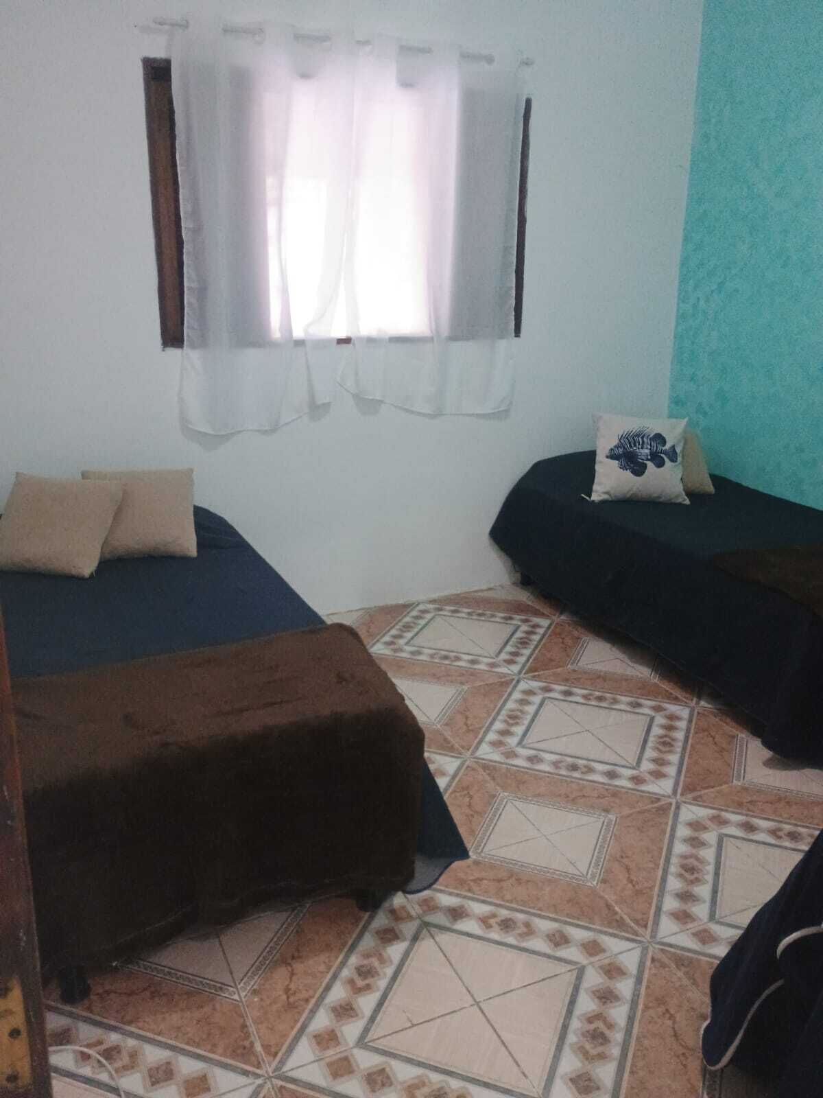 A room with two beds, a window with sheer curtains, a tiled floor, and a teal accent wall.