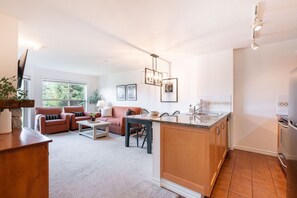 Ein modernes Wohnzimmer mit Sofa, Couchtisch und Essbereich. Es gibt eine Küche mit Spüle und Herd. Der Raum hat große Fenster und eine Deckenleuchte.