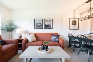 A living room with a sofa, a coffee table, a dining area with chairs, and framed pictures on the wall. - SKI-IN/SKI-OUT 1BDR Condo with Pool & Hot Tub | Spectacular location! (Whistler)