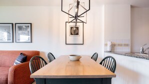 A dining area with a wooden table, chairs, a bowl, framed pictures on the wall, and a kitchen in the background. - SKI-IN/SKI-OUT 1BDR Condo with Pool & Hot Tub | Spectacular location! (Whistler)