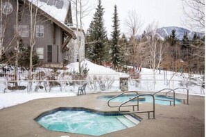 A pool area with two hot tubs, surrounded by a wooden deck and a stone wall. - SKI-IN/SKI-OUT 1BDR Condo with Pool & Hot Tub | Spectacular location! (Whistler)