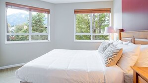 Quarto com uma cama grande, duas janelas com persianas, mesa de cabeceira com luminária e vista para árvores e montanhas.