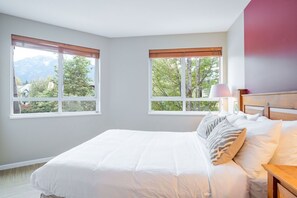 Ein Schlafzimmer mit einem großen Bett, zwei Fenstern mit Jalousien, einem Nachttisch mit Lampe und Blick auf Bäume und Berge.