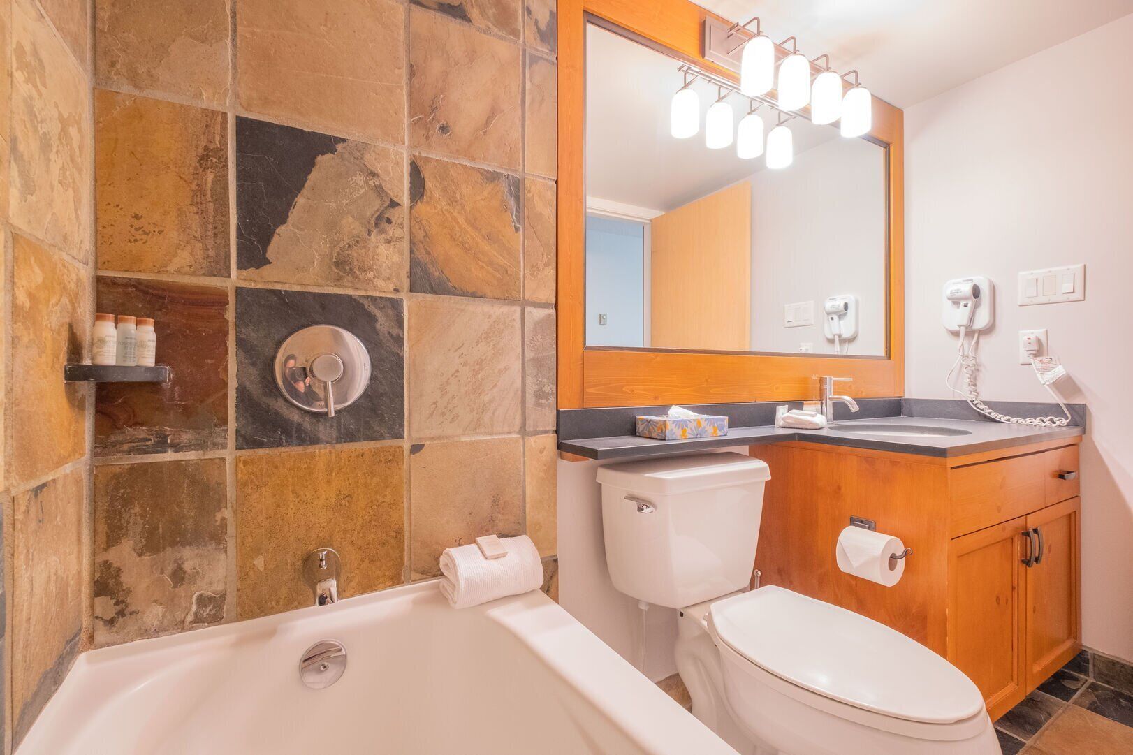 A bathroom with a bathtub, toilet, and sink, featuring stone tile walls and wooden cabinets.