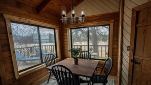 A wooden interior with a dining table, chairs, a chandelier, and large windows.
